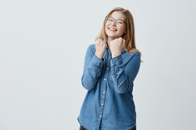 Waist up portrait of delightful smiling happy girl with eyeglasses dressed in denim shirt, clenches fists, rejoices good news, being confident in her big success. I have done it!