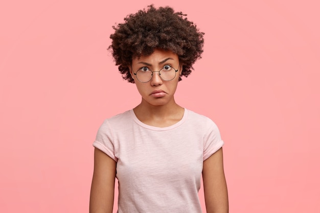 Waist up portrait of dissatisfied African American female with negative angry facial expression, looks in displeasure at someone, wears casual t-shirt, being annoyed spend weekend at home alone