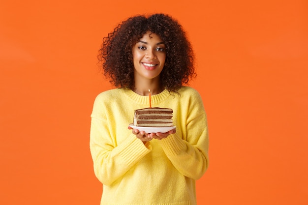 Портрет талии мечтательной афро-американской девушки с афро-стрижкой, держащей тарелку с праздничным тортом и зажженной свечой, задувающей его, чтобы загадать желание, счастливо улыбаясь, празднуя над оранжевой стеной.