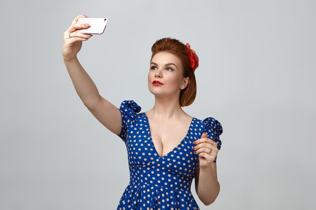 Waist up portrait of gorgeous fashionable young lady dressed like 1950s pin up girl holding smart phone above her and taking selfie