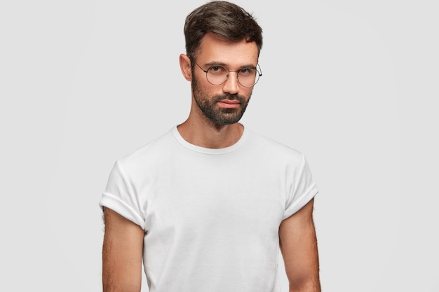 심각한 표정으로 잘 생긴 수염 난 남성의 초상화를 허리 위로 올리고 무언가에 대해 고민하고 둥근 안경과 흰색 캐주얼 티셔츠를 입고 실내 포즈를 취합니다. 사람과 표정