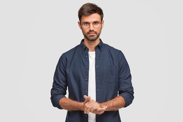 Waist up portrait of handsome serious unshaven male keeps hands together, dressed in dark blue shirt, has talk with interlocutor, stands against white wall. Self confident man freelancer
