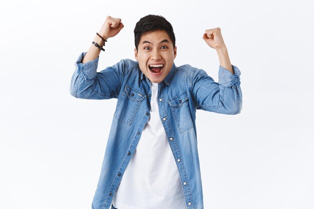 waist-up portrait of happy, excited young smiling man shouting hooray or yes, raising hands up, fist pump in joy, rejoicing over win, achieve goal, celebrate victory or success, white wall