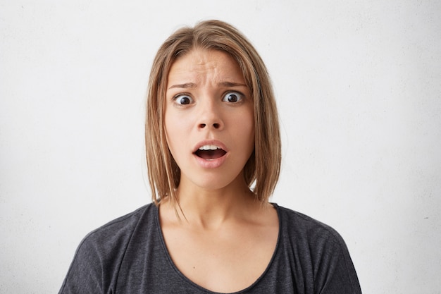 Free photo waist up portrait of shocked woman with trendy hairstyle staring with big dark eyes and widely opened mouth being in panic