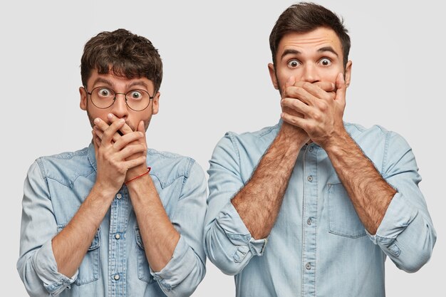 Waist up portrait of shocked young men cover mouth with both hands, stare with unexpected look, notice something unbelievable, dressed in denim shirts, isolated over white wall