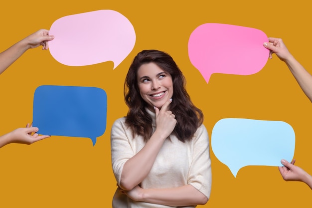 Waist-up portrait of a smiling cute lady pondering over something surrounded by hands with four question and answer signs