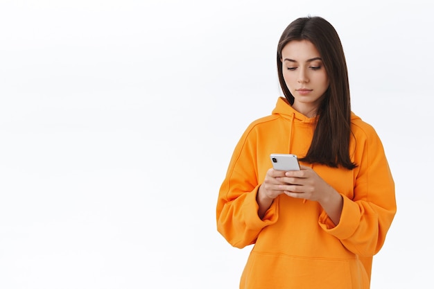 휴대 전화를 사용하여 주황색 후드티를 입은 젊은 힙스터 소녀의 허리 위로 초상화, 진지한 얼굴로 스마트폰 디스플레이 보기, 메시지 쓰기, 친구에게 문자 보내기 또는 온라인에 게시할 사진 편집