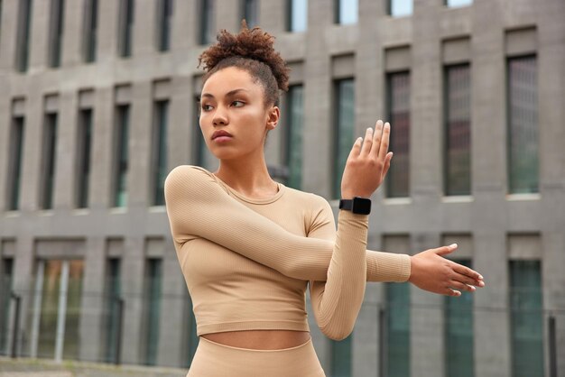 Waist up shot of active slim woman stretches arms before workout dressed in top has curly combed hair concentrated away poses against blurred building background prepares for cardio training
