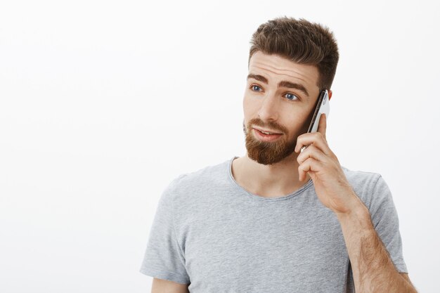 Waist-up shot of ambitious good-looking busy man with beard, moustache and blue eyes looking left serious and determined discussing business via smartphone holding cellphone near ear against grey wall