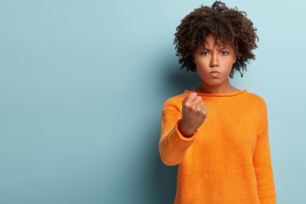 Foto gratuita mezzo busto di donna infastidita con taglio di capelli afro, mostra il pugno, guarda con rabbia, minaccia di vendetta, indossa un maglione arancione casual, isolato su un muro blu con spazio vuoto. ascoltami