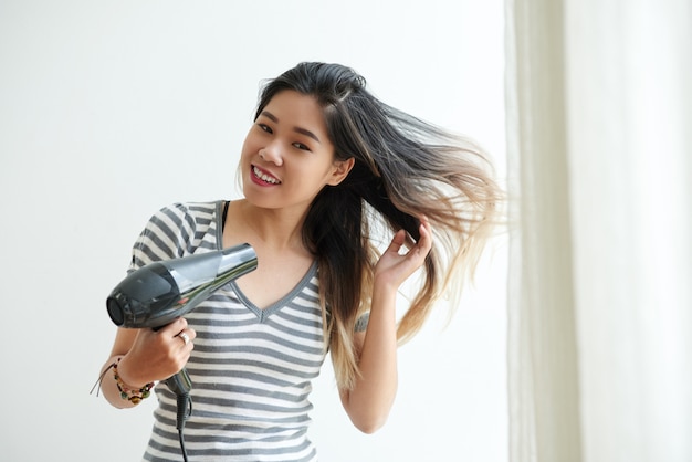 Free photo waist up shot of asian girl blow-drying her hair at home