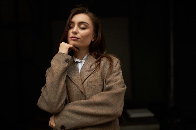 Waist up shot of attractive fashionable young European female with long brown hair posing isolated keeping eyes closed, having dreamy peaceful facial expression, wearing mens jacket, smiling