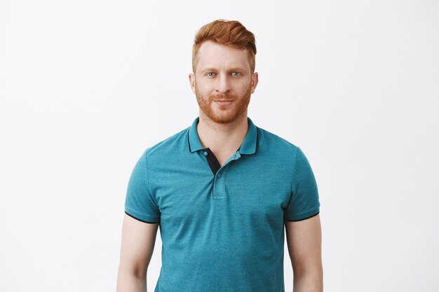 Снимок талии привлекательного мужественного мужчины с рыжими волосами в зеленой рубашке-поло, ухмыляющегося и смотрящего с уверенным и самоуверенным выражением лица, чувствуя себя спокойно