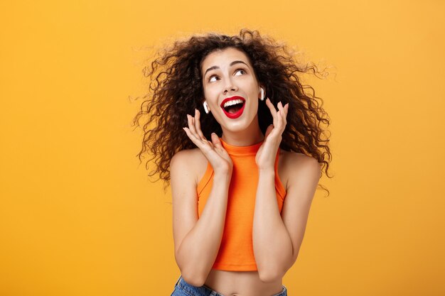 Waist-up shot of attractive silly curly-haired european female in cropped top using wireless earphones touching earbuds and gazing at upper right corner delighted and carefree over orange wall.