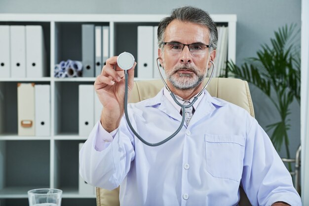 Waist up shot of bearded senior doctor with stethoscope turned to camera