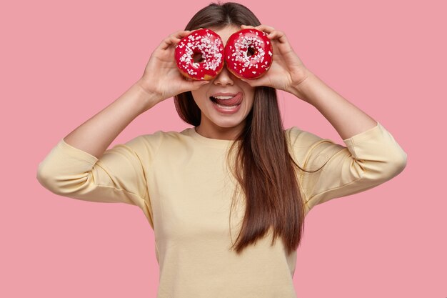 Waist up shot of beautiful young woman shows tongue from pleasant taste, covers eyes with doughnuts, has fun indoor, dressed in yellow clothes