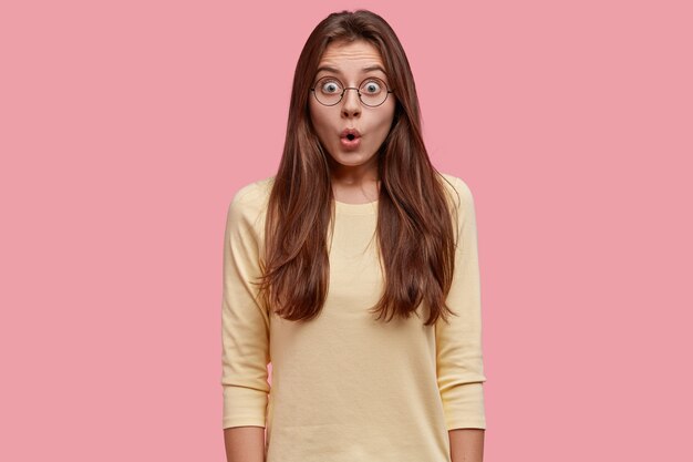 Waist up shot of brunette young European woman amazed by shocking rumors, gasps from shock, wears yellow sweater