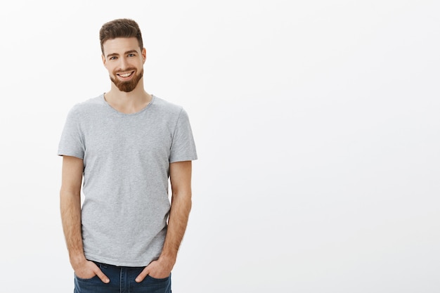 Waist-up shot of carefree ambitious and handsome bearded male with moustache and blue eyes holding hands in pockets of jeans smiling with perfect white teeth with assured expression over gray wall