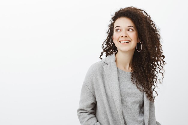 Waist-up shot of carefree happy european female entrepreneur in earrings and stylish coat, gazing left with broad joyful smile