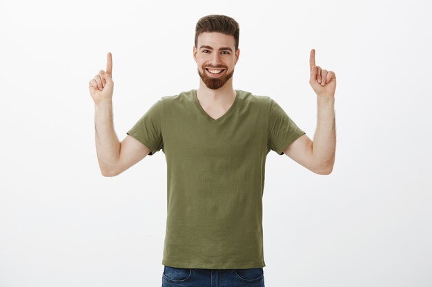 Waist-up shot of charismatic caucasian male with beard in olive t-shirt raising hands and pointing up with index fingers. smiling pleased and delighted with happy expression over white wall