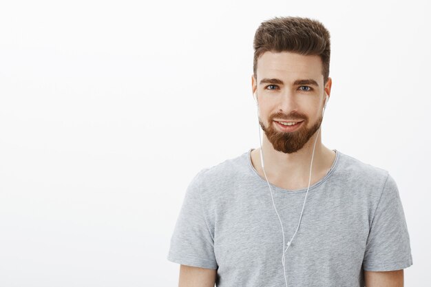 Waist-up shot of charismatic handsome adult man with beard and blue eyes smiling pleased and daring wearing earphones searching right mood for creative ideas while listening music over gray wall