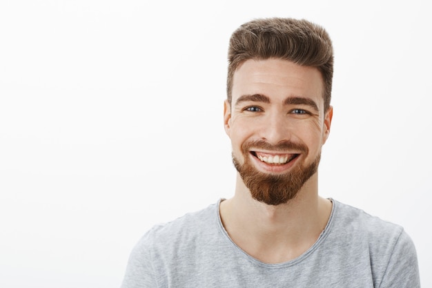 Waist-up shot of charming carefree and optimistic handsome man with beard, moustache and cute blue eyes laughing joyfully squinting from joy and amusement having fun against gray wall