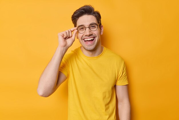 Waist up shot of cheerful brunet guy keeps hand on rim of spectacles wears casual t shirt and round spectacles being in good mood poses against vivid yellow background Happy emotions concept