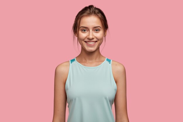 Waist up shot of cheerful Caucasian woman with appealing appearance, dressed in casual vest, satisfied with sport training, model against pink wall. Facial expressions and emotions concept