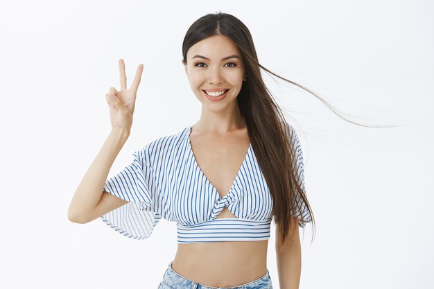 Waist-up shot of chill charming european female with perfect body and long dark hair in cropped top showing peace gesture