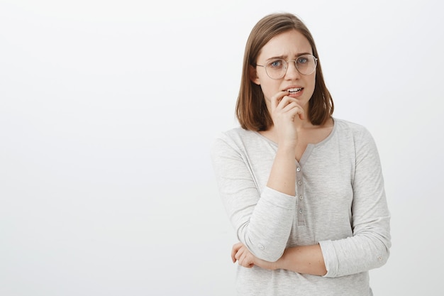 Waist-up shot of concerned troubled cute european female in glasses and blouse holding finger on lip frowning looking nervous and worried waiting for news from hospital holding hand crossed on body