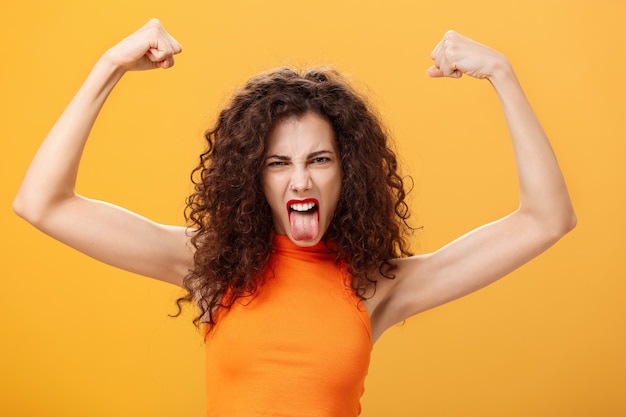 Foto gratuita colpo in vita di una donna caucasica fresca e audace in top arancione con un tatuaggio sul braccio accigliato che fa una faccia buffa che sporge la lingua alzando le mani mostrando i muscoli che sentono potere e forza. copia spazio