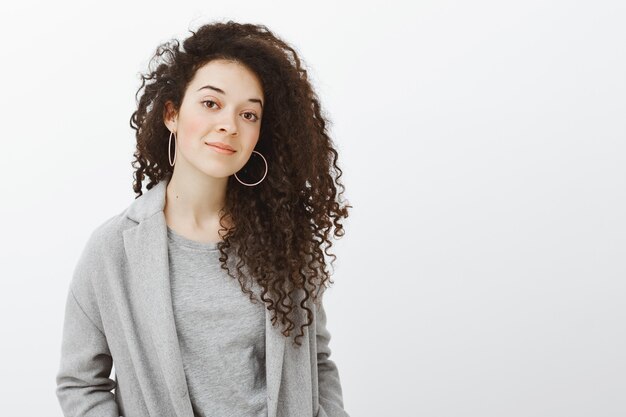 Waist-up shot of creative beautiful woman with curly hair in grey coat and earrings, smiling broadly and tilting head, listening carefully while working with coworkers