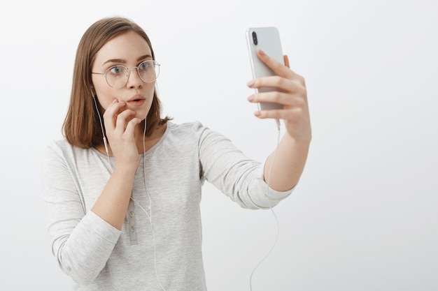 Waist-up shot of creative curious and entertained charming woman in glasses wearing earphones raising hand with smartphone gazing intrigued and interested at device screen watching amazing video