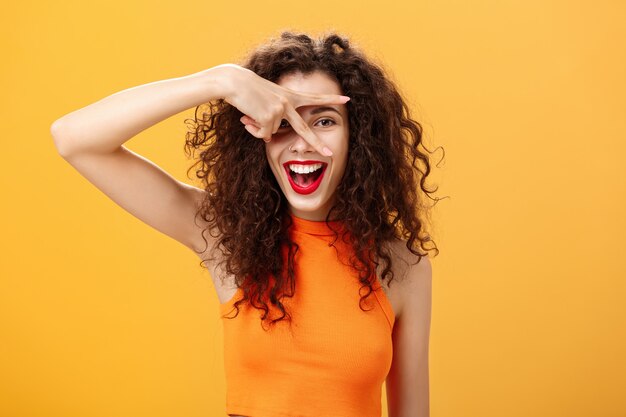 Waist-up shot of creative joyful caucasian female with curly hairstyle and small tattoo showing peace gesture and peeking through fingers at camera happily smiling broadly having fun over orange wall.