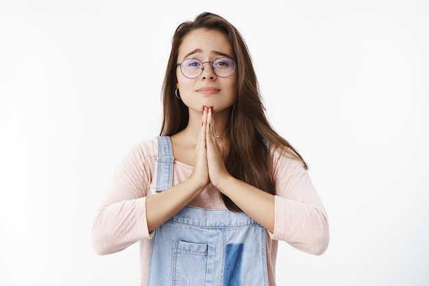 Waist-up shot of cute charming female brunette in glasses smiling and holding hands in pray raising eyebrows questioned as begging and waiting for positive answer while asking help over gray wall