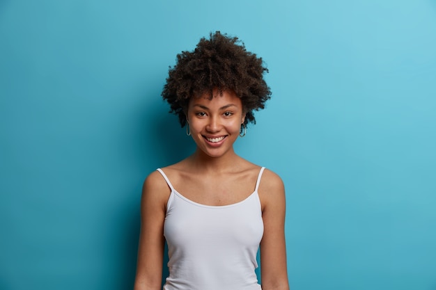 Снимок талии темнокожей молодой женщины с афро-прической, приятно улыбается, одетая в повседневный белый жилет, приятно разговаривает, изолирована на синей стене, выражает счастье и нежность