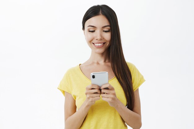 Снимок талии довольной беззаботной привлекательной женщины-коллеги в желтой модной футболке, держащей смартфон и смотрящей на экран телефона