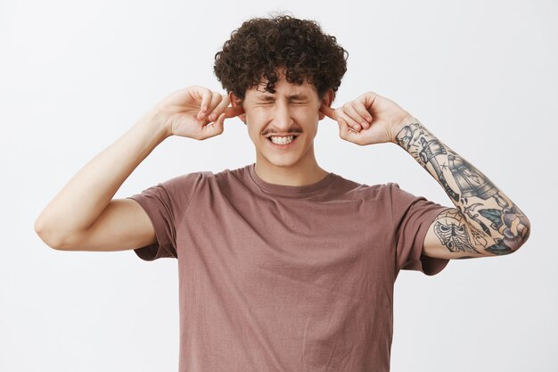 Waist-up shot of displeased intense modern hipster male student with moustache curly hair and tattooed arm clenching teeth from discomfort covering ears with index fingers not hear annoying loud sound