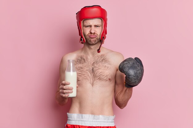 Waist up shot of displeased topless sportsman holds glass bottle of milk feels tired of training and going in for sport wears boxing gloves protective helmet on head