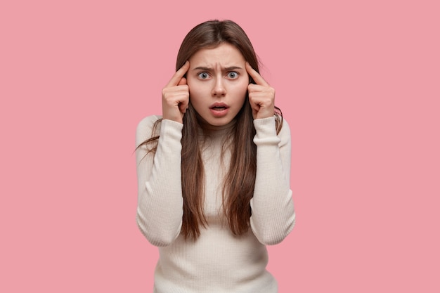 Waist up shot of dissatisfied dark haired woman has stupefied facial expression, keeps index fingers on temples, frowns face, has long black hair