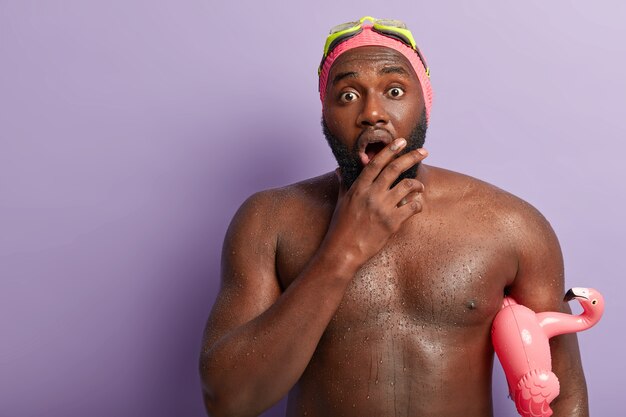 Waist up shot of embarrassed dark skinned guy opens mouth widely, has muscular body, swims in swimming pool