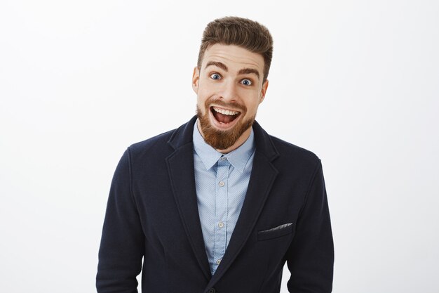 Waist-up shot of enthusiastic delighted and surprised charismatic young rich bachelor in stylish suit with beard and blue eyes smiling broadly from amazement and joy gazing impressed