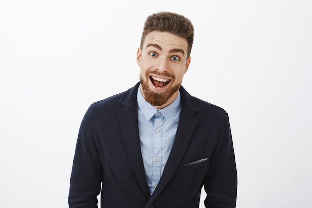 Waist-up shot of enthusiastic delighted and surprised charismatic young rich bachelor in stylish suit with beard and blue eyes smiling broadly from amazement and joy gazing impressed