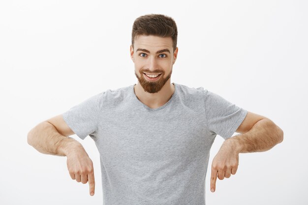 Waist-up shot of excited and confident charming brunet man with beard and moustache pointing down and smiling broadly looking with enthusiasm and excited expression against grey wall