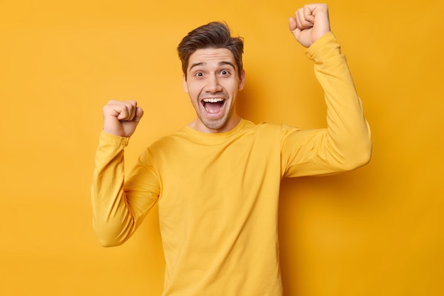 Waist up shot of excited handsome man makes triumph gesture shakes arms fist bump exclaims loudly dressed in casual jumper isolated over vivid yellow background filled with joy achieved goal