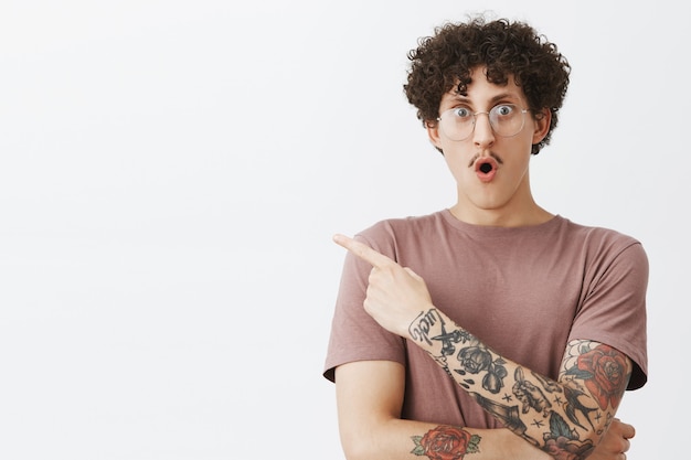 Waist-up shot of excited and impressed emotive attractive male student with stylish moustache curly dark hair and tattooed arm gasping folding lips from amazement and pointing at upper left corner
