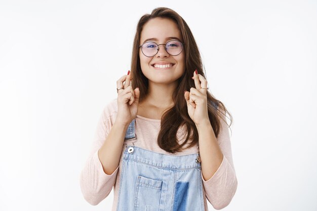 Waist-up shot of excited smiling hopeful cute woman in glasses biting lower lip from excitement and joy as waiting for good news with crossed fingers