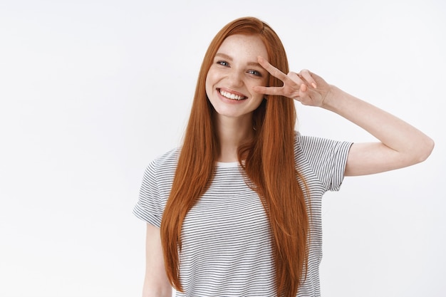 Waist-up shot friendly kind optimistic attractive young lucky teenage redhead girl blue eyes tilting head smiling white teeth show victory peace gesture enjoy party, standing white wall