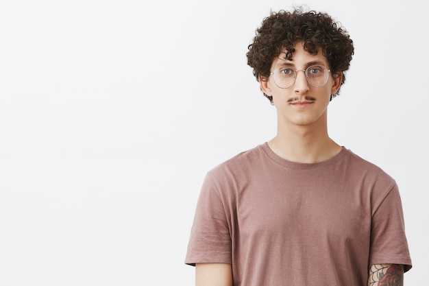 Free photo waist-up shot of friendly-looking calm and carefree young man with moustache and curly hairstyle wearing glasses and brown t-shirt smiling cute standing casually over gray wall