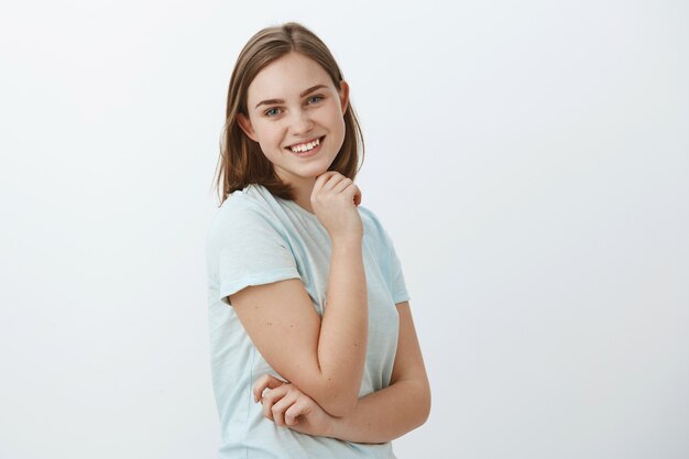 Waist-up shot of friendly-looking joyful lucky girl with brown short haircut standing in profile holding hand on cheek and turning with broad happy smile posing against grey wall